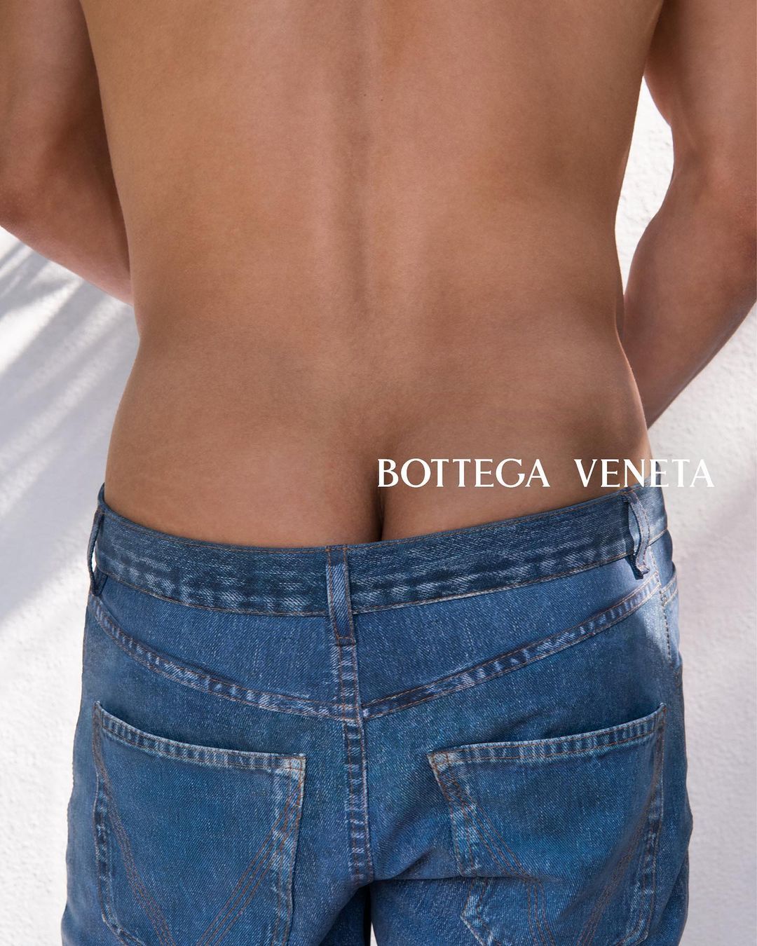 Rear view of a shirtless man wearing blue jeans, showcasing a fashion advertisement with 'BOTTEGA VENETA' text against a light backdrop.