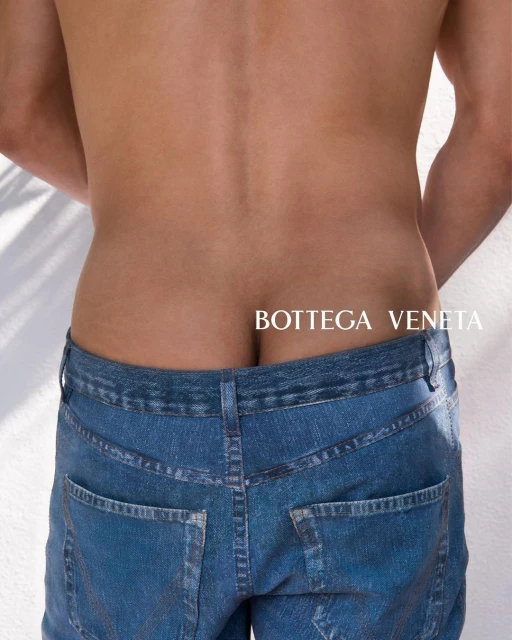 Rear view of a shirtless man wearing blue jeans, showcasing a fashion advertisement with 'BOTTEGA VENETA' text against a light backdrop.