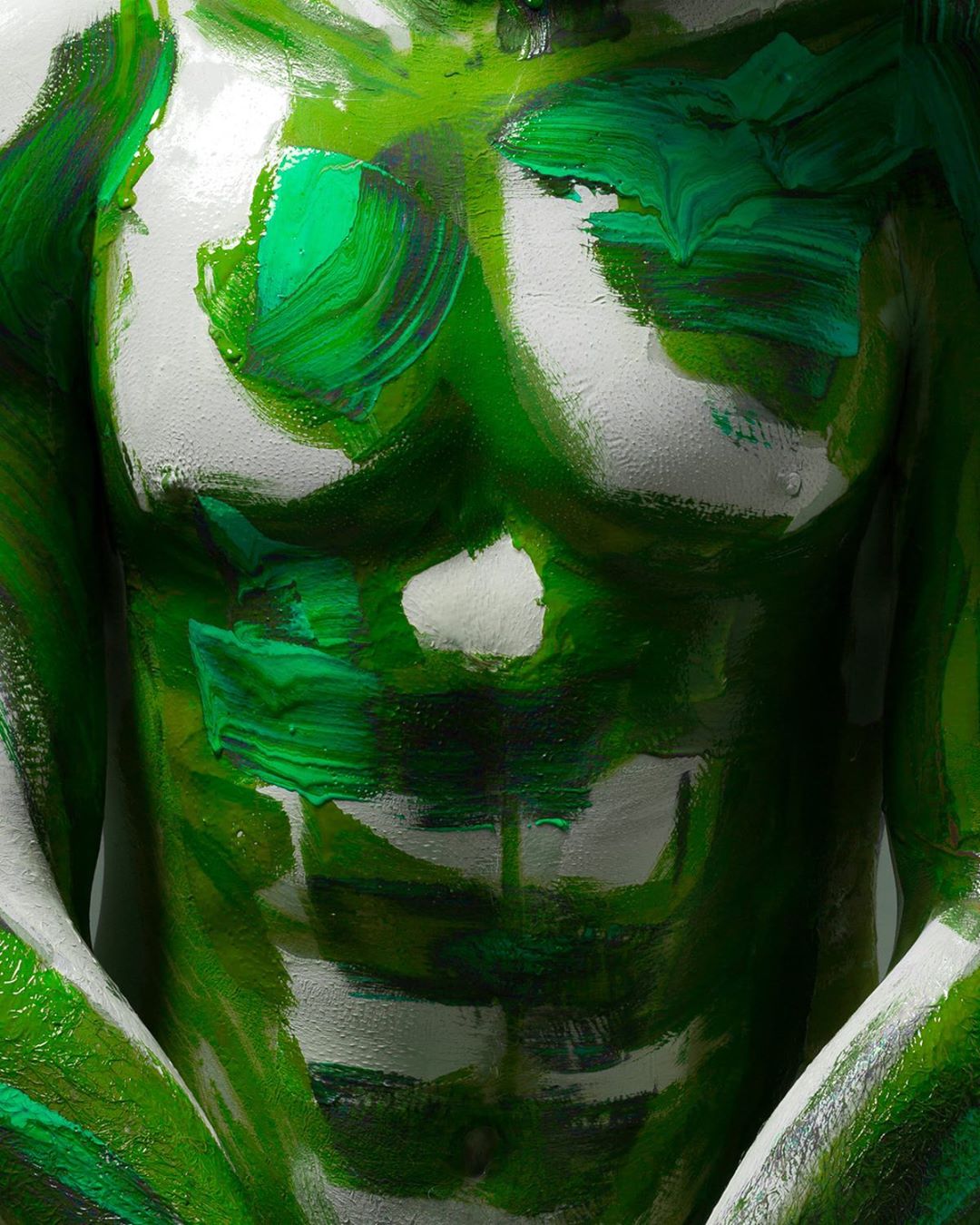 Male torso covered in shades of green paint against a white backdrop. Artful body painting, showcasing artistic design and vivid colours.