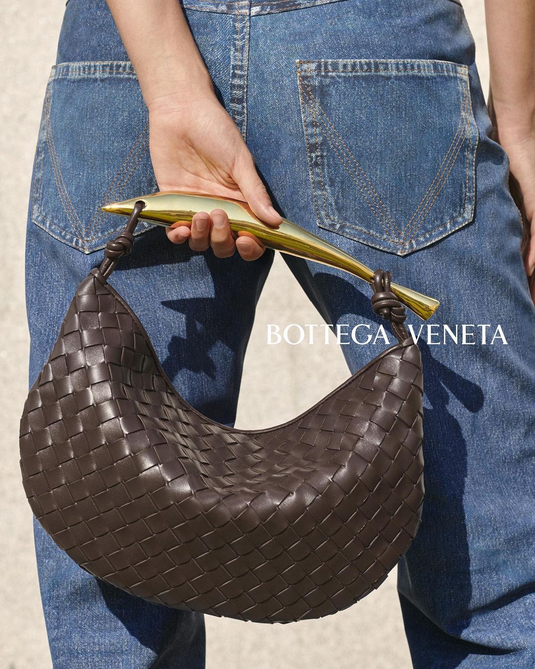 Woven brown leather hobo bag with a golden metal handle, held by a person wearing denim jeans against a neutral background.