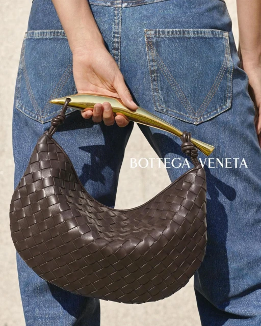 Woven brown leather hobo bag with a golden metal handle, held by a person wearing denim jeans against a neutral background.