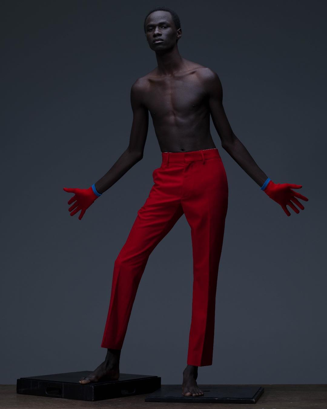 An African male model with bare torso wearing red pants and gloves stands on small platforms against a grey background in a studio shot.