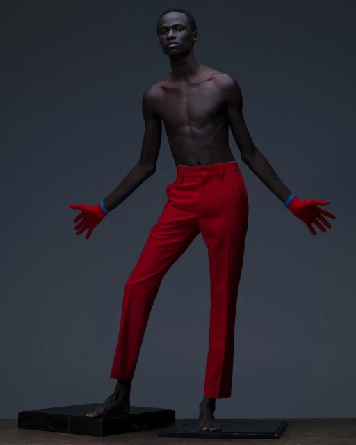 An African male model with bare torso wearing red pants and gloves stands on small platforms against a grey background in a studio shot.