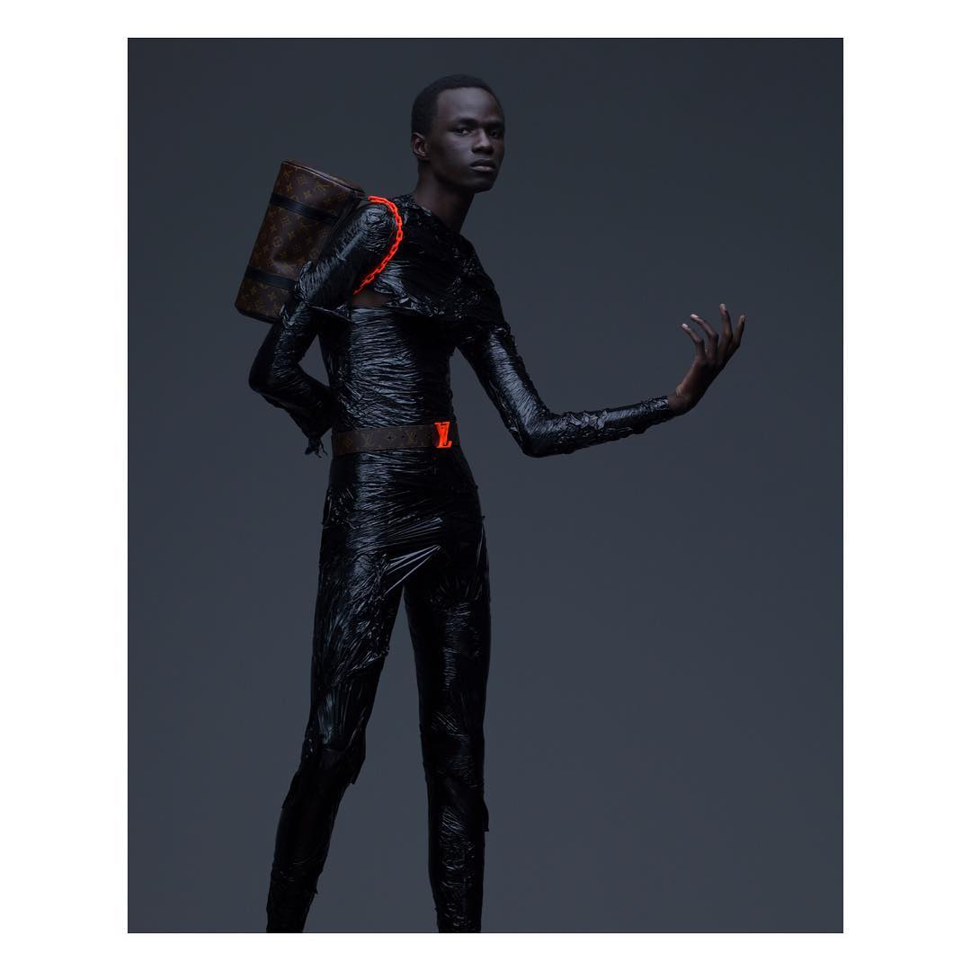 Male fashion model in dark clothing with texture and a Louis Vuitton satchel poses in a studio with a dark background and neutral lighting.