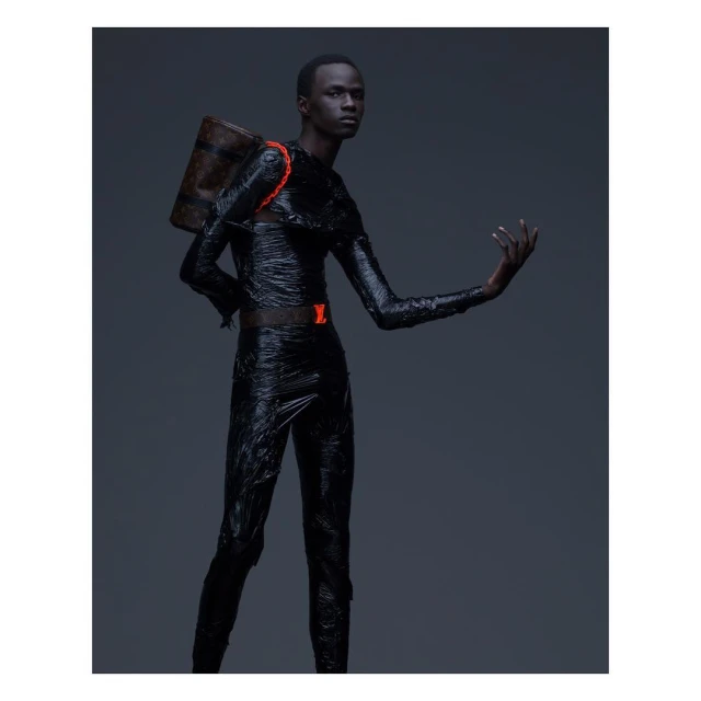 Male fashion model in dark clothing with texture and a Louis Vuitton satchel poses in a studio with a dark background and neutral lighting.