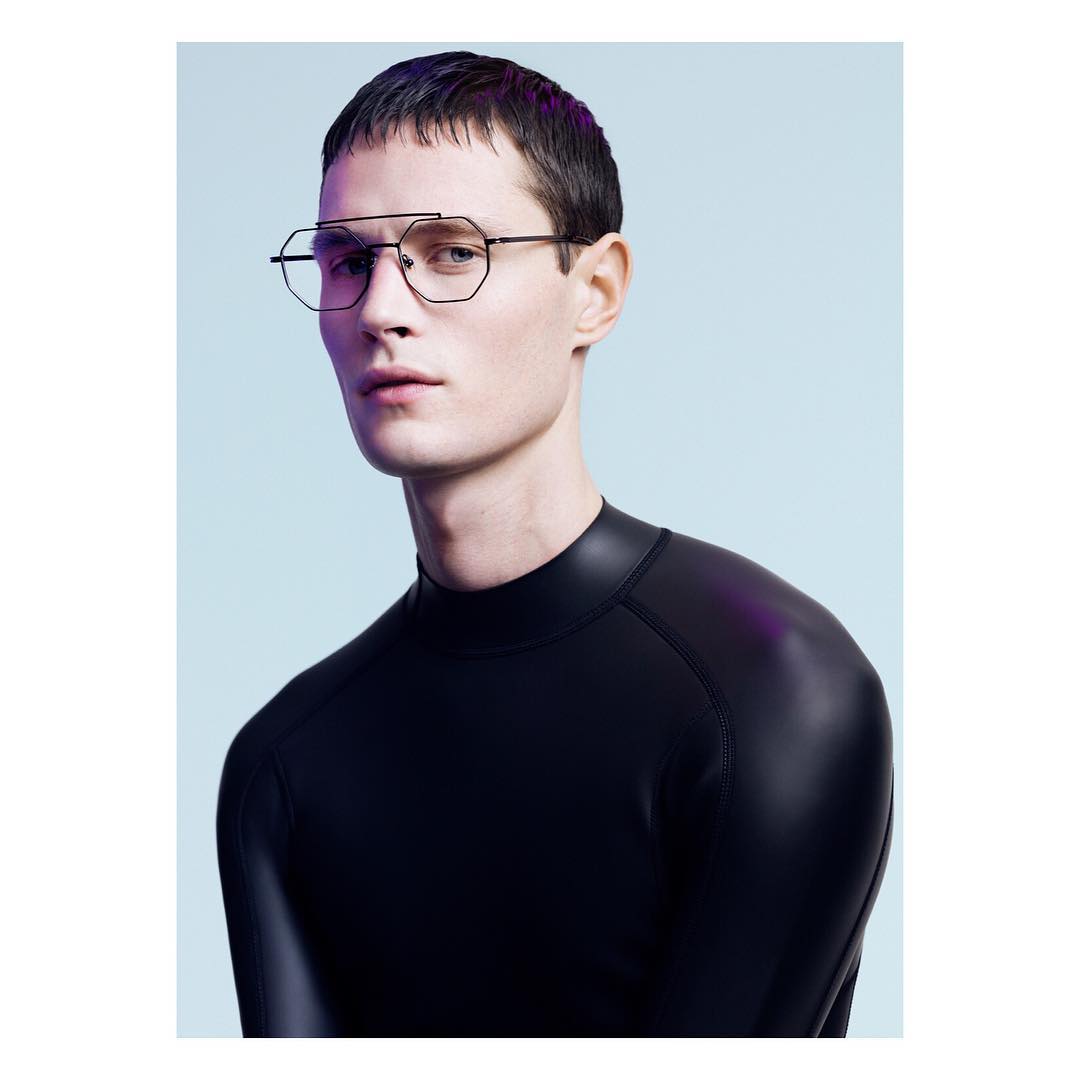Close-up studio portrait of a young adult male model with short dark hair wearing geometric eyeglasses and a black mock turtleneck shirt.