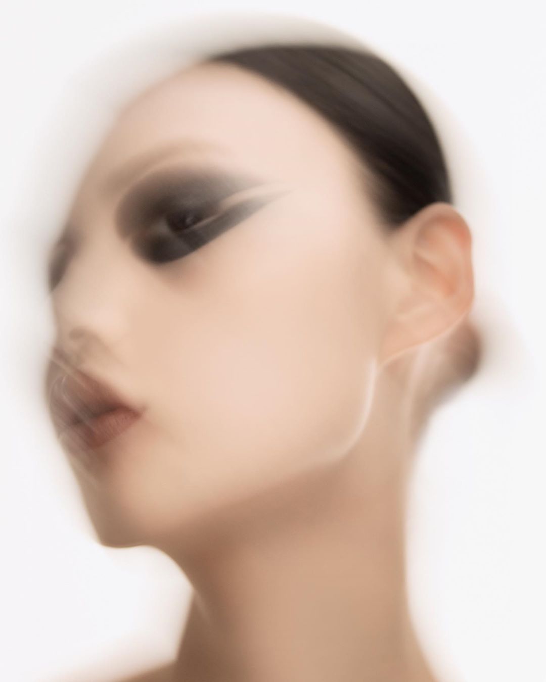 Close-up beauty shot of an Asian woman with black eye makeup, slicked-back dark hair, and fair skin against a diffused white background.