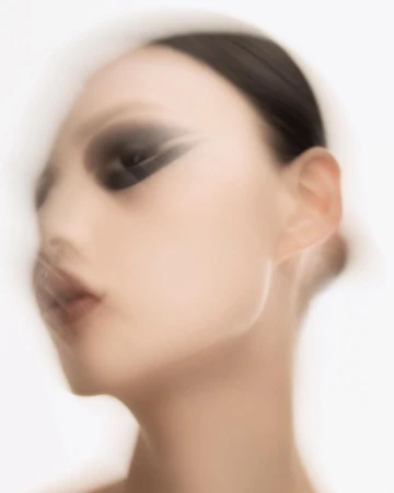 Close-up beauty shot of an Asian woman with black eye makeup, slicked-back dark hair, and fair skin against a diffused white background.