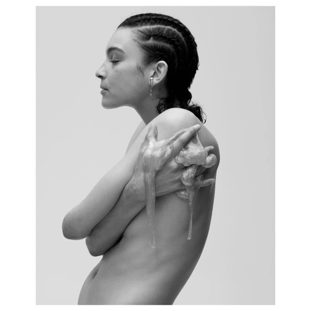 Black and white portrait of a woman with bare shoulders, silver paint on her hands, and braided hair in front of a neutral background.