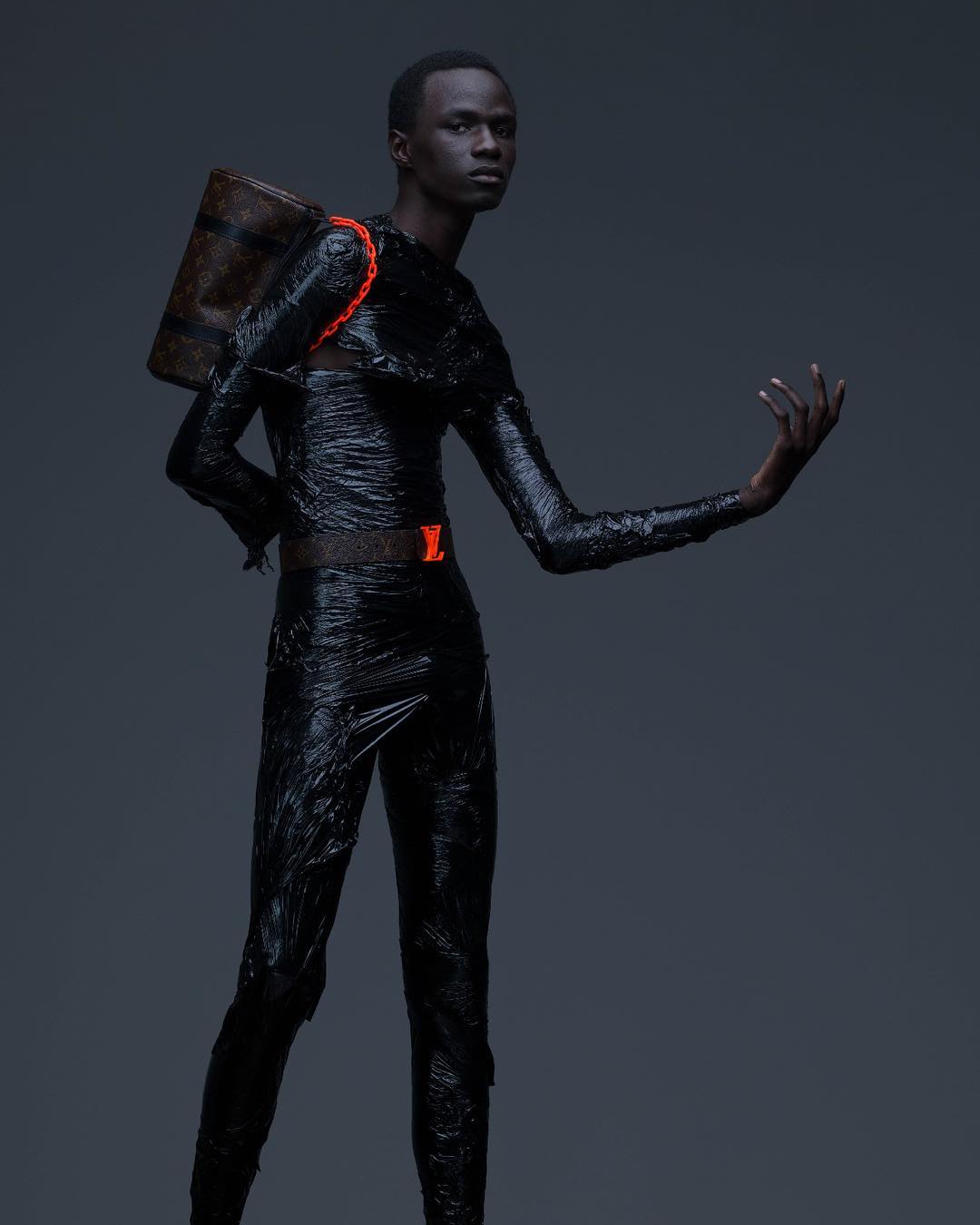 Fashionable adult male poses in black, shiny outfit with Louis Vuitton backpack against a dark gray background in a studio shot.