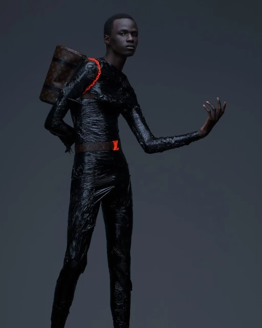 Fashionable adult male poses in black, shiny outfit with Louis Vuitton backpack against a dark gray background in a studio shot.