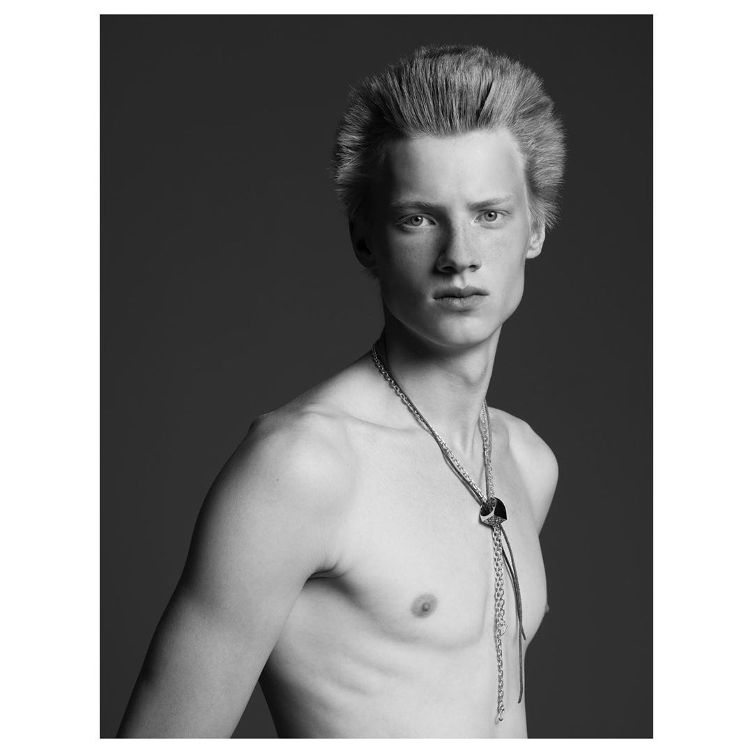 Black and white portrait of a shirtless young adult male with a bolo tie, posing in a photo studio setting with a neutral expression.