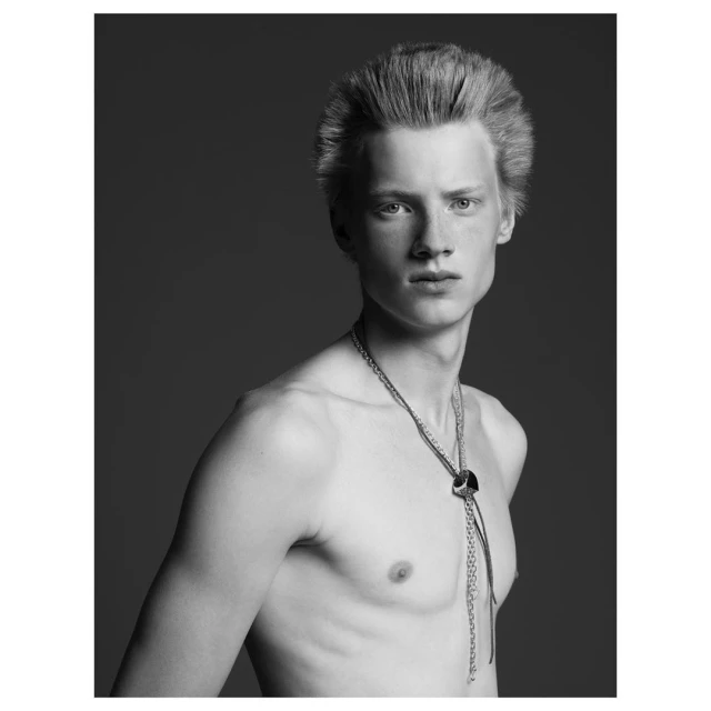 Black and white portrait of a shirtless young adult male with a bolo tie, posing in a photo studio setting with a neutral expression.