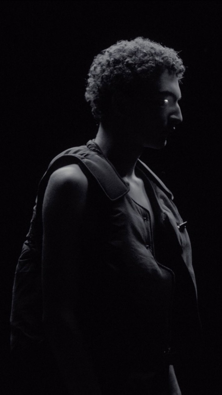 Monochromatic portrait of adult male with curly hair in front of black background wearing a vest, illuminated to emphasize silhouette.