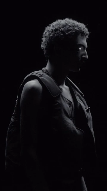 Monochromatic portrait of adult male with curly hair in front of black background wearing a vest, illuminated to emphasize silhouette.