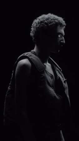Monochromatic portrait of adult male with curly hair in front of black background wearing a vest, illuminated to emphasize silhouette.