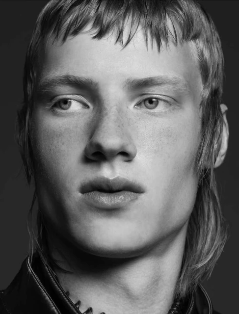 Black-and-white close-up studio portrait of a fair-skinned young adult male with a mullet and dark leather jacket, looking left.