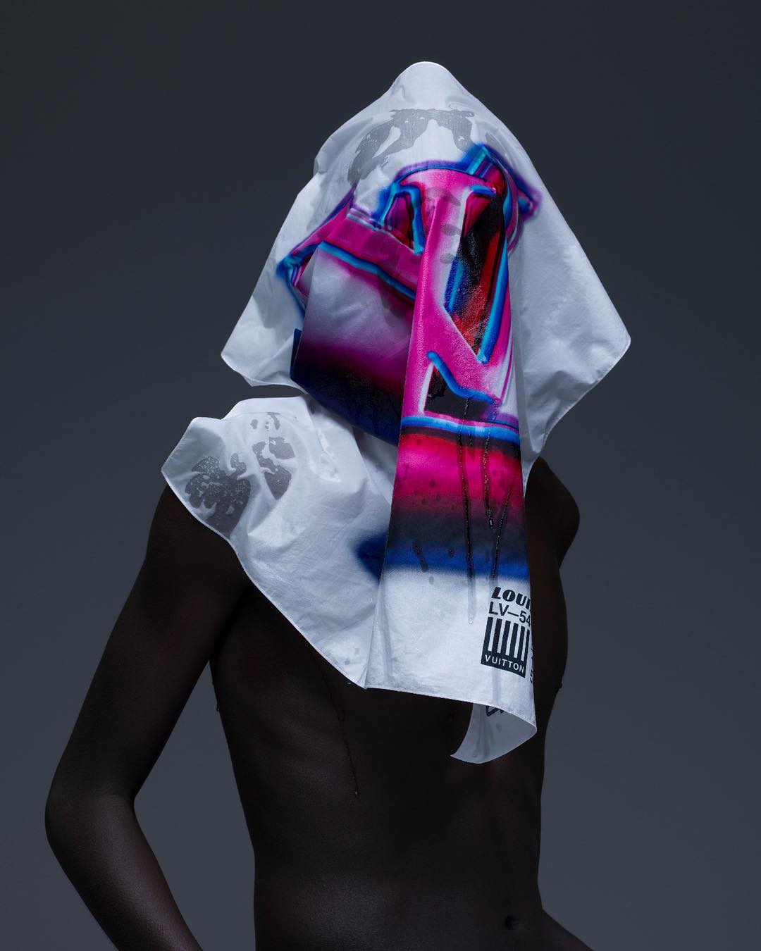 Person draped in a white Louis Vuitton printed textile, back to the camera in a studio setting with neutral grey background.
