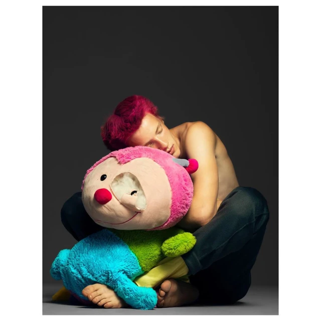 A young adult with magenta hair is hugging a large, colorful stuffed toy against a dark grey background in an indoor setting.