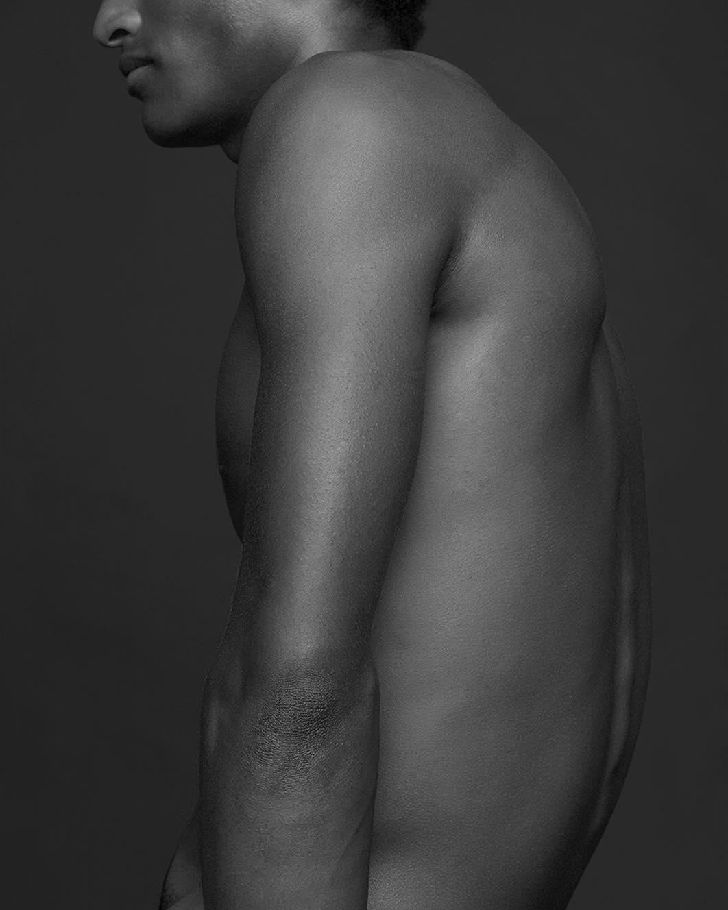 Monochrome portrait of a bare-chested, dark-skinned male torso against a dark gray background, featuring prominent back muscles.