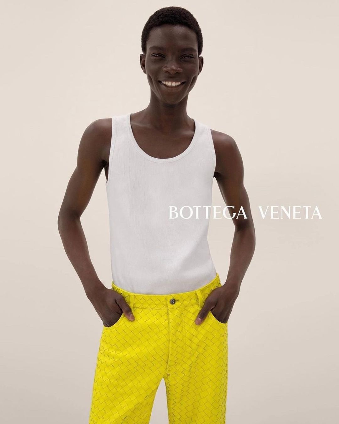 Fashion shot of an adult male model with a smile, wearing a white tank top and yellow textured pants, posing indoors in a photo studio.