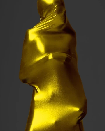 Figure draped in shimmering gold fabric creating abstract human form with dramatic highlights and shadows against dark gray background.
