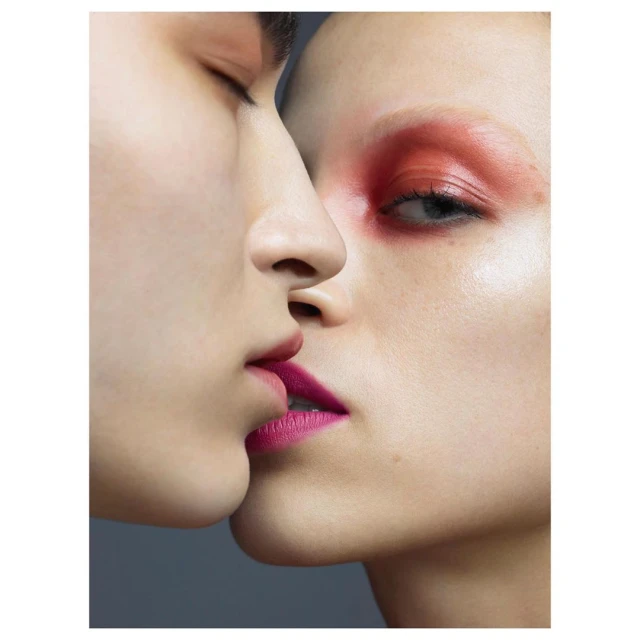 Close-up of two faces with makeup: bright pink lipstick, bold red eyeshadow on fair skin, against a dark gray background in studio lighting.