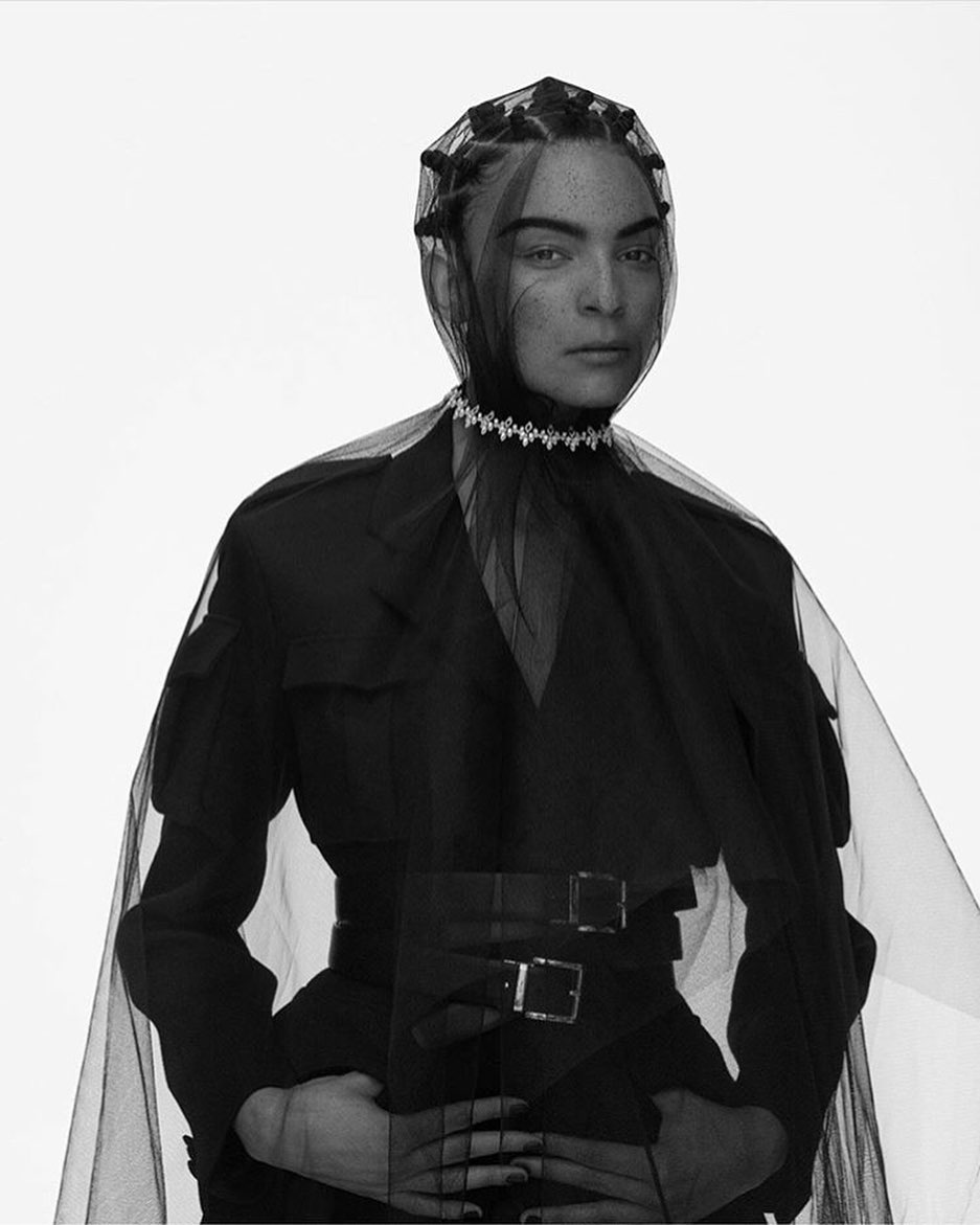 Monochrome portrait of a female model wearing a black jacket, a net headpiece, and a jeweled collar, set against a bright white backdrop.