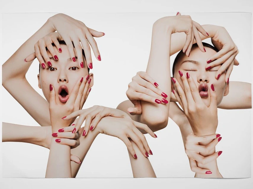 Two Asian women frame their faces with hands in a beauty shot, displaying red manicured nails against a clean white background.