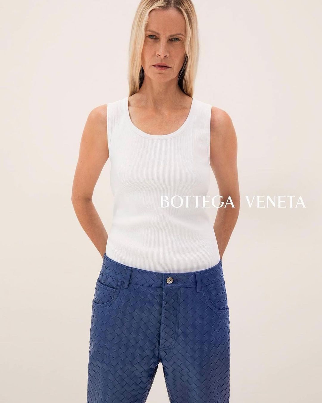 Blonde woman in a white tank top and woven blue pants stands in a brightly lit neutral studio, showcasing a modern, high-fashion look.
