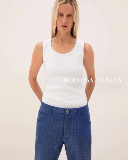 Blonde woman in a white tank top and woven blue pants stands in a brightly lit neutral studio, showcasing a modern, high-fashion look.