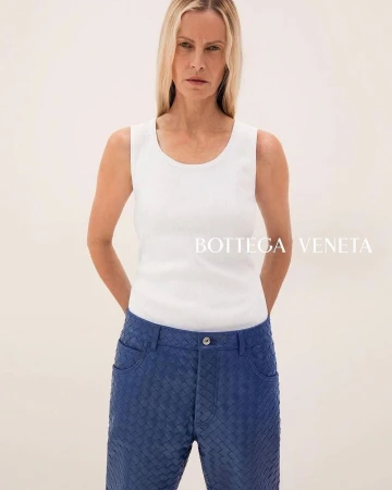 Blonde woman in a white tank top and woven blue pants stands in a brightly lit neutral studio, showcasing a modern, high-fashion look.