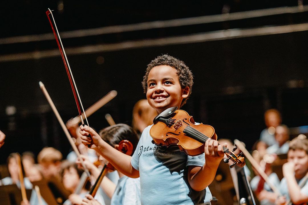 Young musician performs with philharmonic orchestra, smiling with violin and bow in hand, surrounded by fellow musicians.