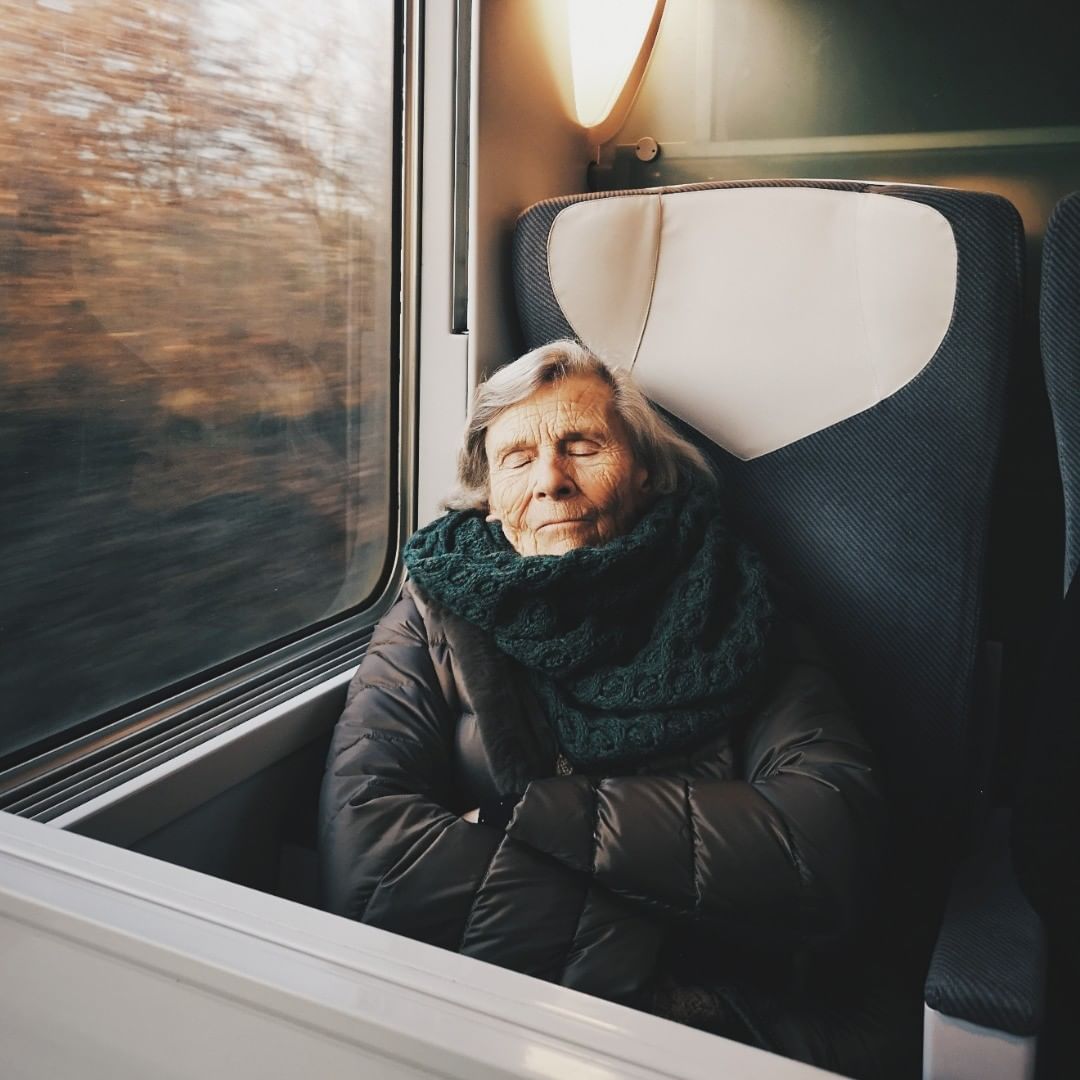 Elderly woman rests peacefully on a train, wearing a green scarf and brown jacket, soft lighting, blurred motion outside the window.