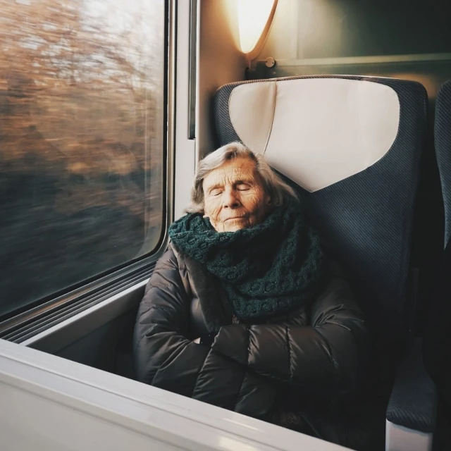 Elderly woman rests peacefully on a train, wearing a green scarf and brown jacket, soft lighting, blurred motion outside the window.