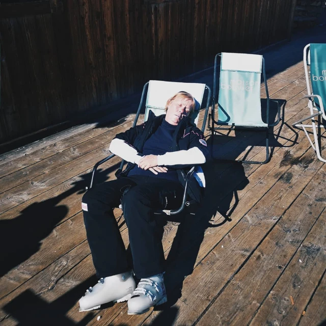 A man in ski boots relaxes on a deck chair under the sun on a wooden deck. There are other chairs in the background.