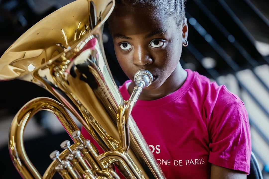 Young musician plays a shiny brass euphonium in an indoor space, wearing a pink shirt, focused, and immersed in the musical performance.