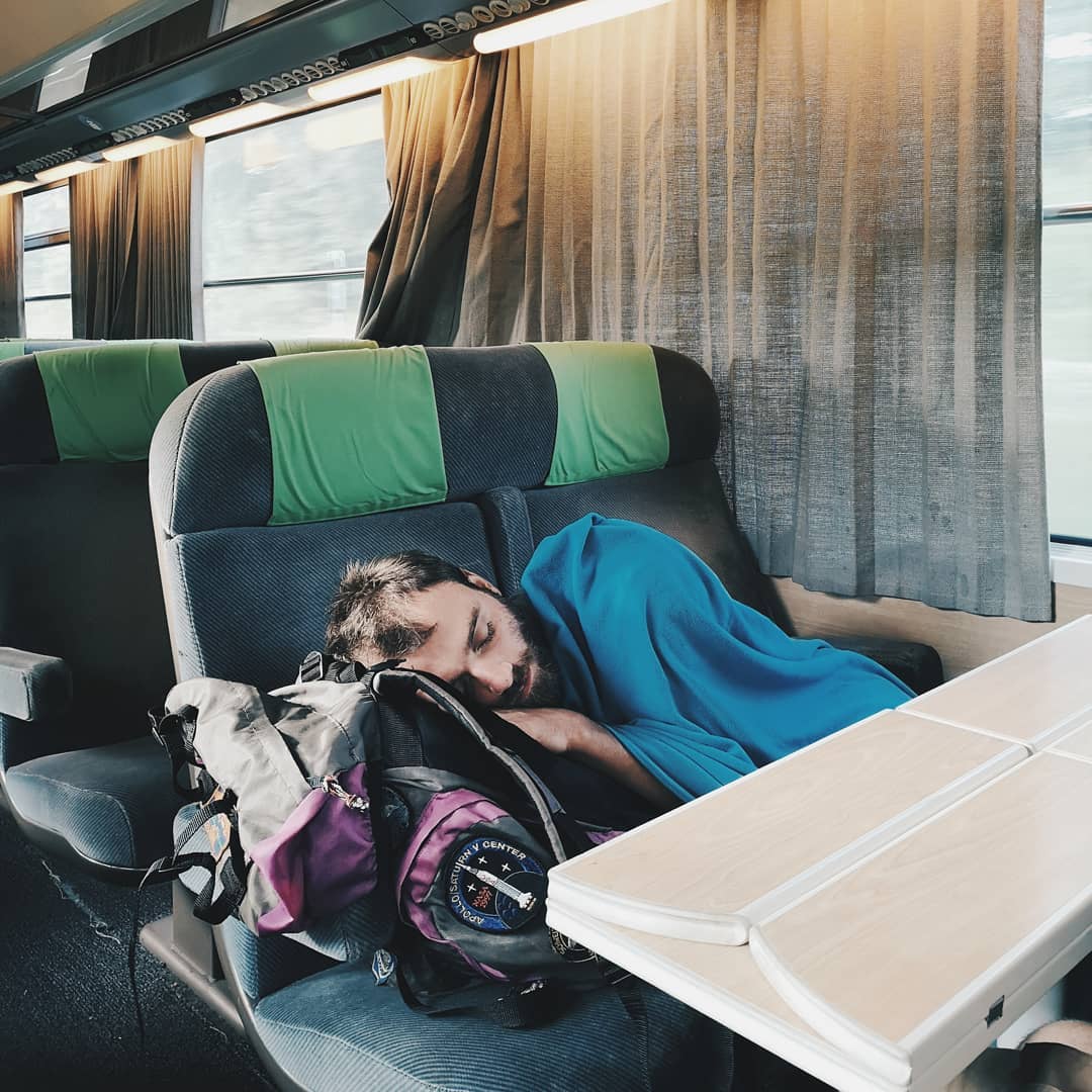 Man sleeping on a train seat with a backpack beside him, next to a table and a window with curtains, creating a tranquil travel scene.