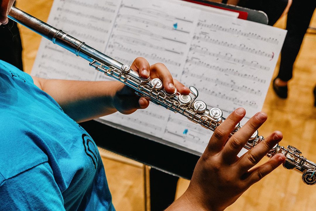 Close-up of a person playing a silver flute with sheet music visible in the background, focusing on the instrument and performer's hands.