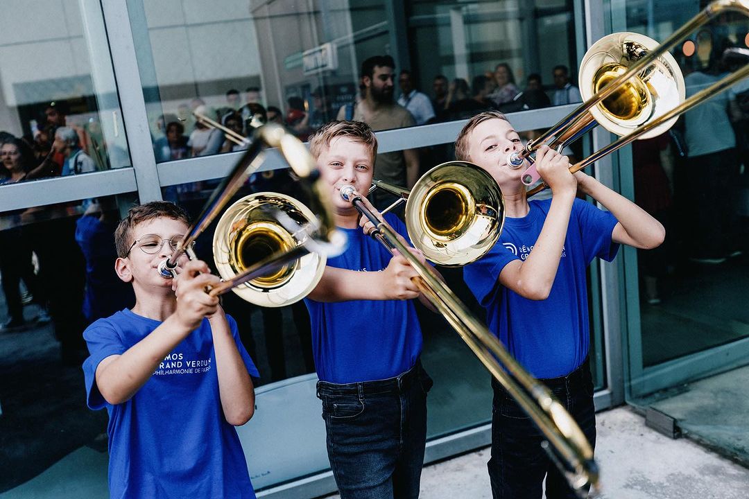 Three young boys in blue shirts play trombones outdoors, surrounded by a crowd, in front of a glass building with natural lighting.