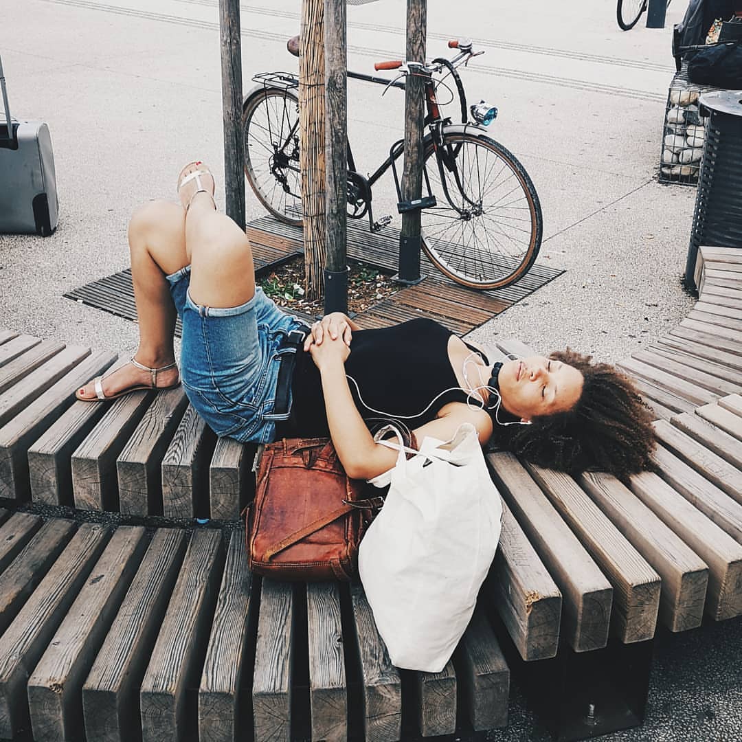 A woman relaxes on a curved wooden bench with her belongings next to her, a bike propped on a tree behind her on an outdoor day.
