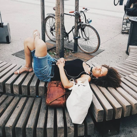 A woman relaxes on a curved wooden bench with her belongings next to her, a bike propped on a tree behind her on an outdoor day.