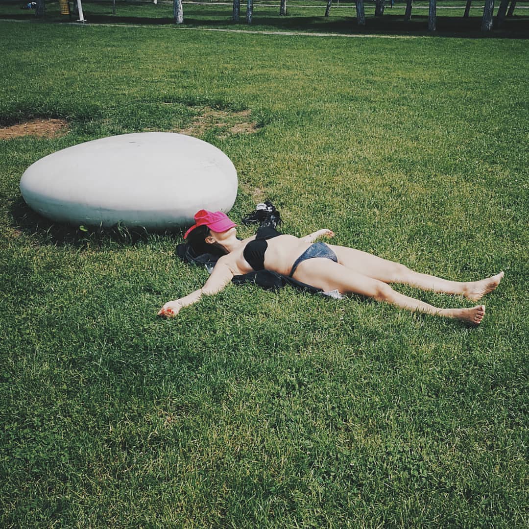 Woman in bikini and pink cap lays on grass near a white oval stone in a park setting, sunbathing on a summer day outdoors.