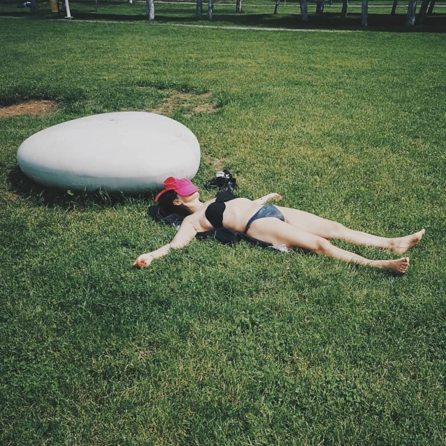 Woman in bikini and pink cap lays on grass near a white oval stone in a park setting, sunbathing on a summer day outdoors.