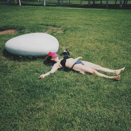 Woman in bikini and pink cap lays on grass near a white oval stone in a park setting, sunbathing on a summer day outdoors.