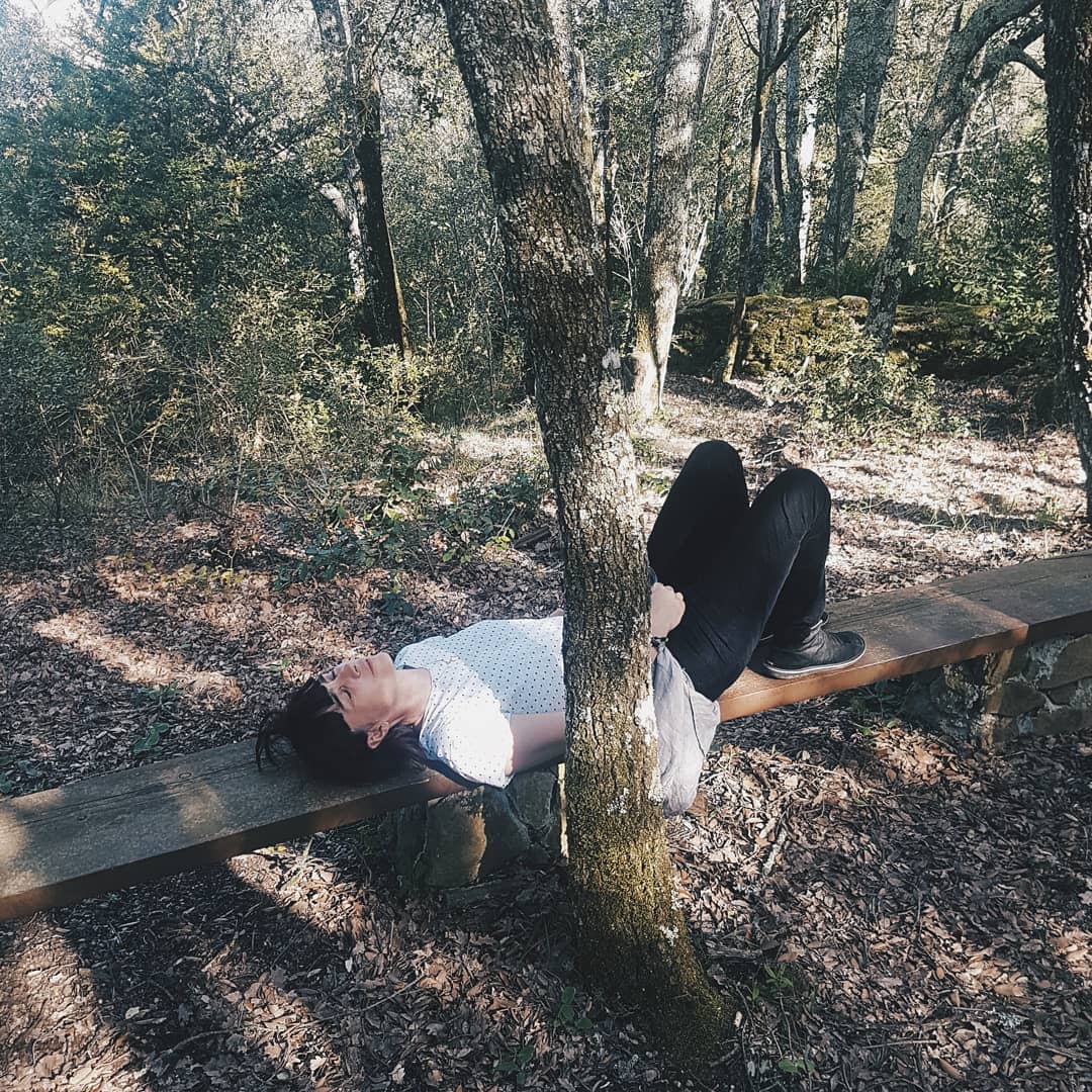 Woman relaxing on a wooden bench in a forest setting, with trees surrounding her. She wears dark pants and a light patterned top.