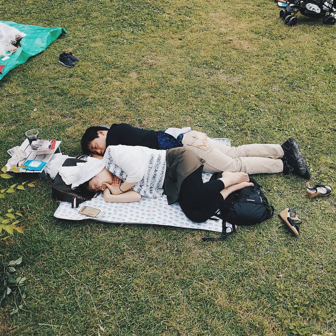 Two people sleeping on a blanket in a grassy outdoor space, with a camping table, shoes, and a stroller visible in the background.