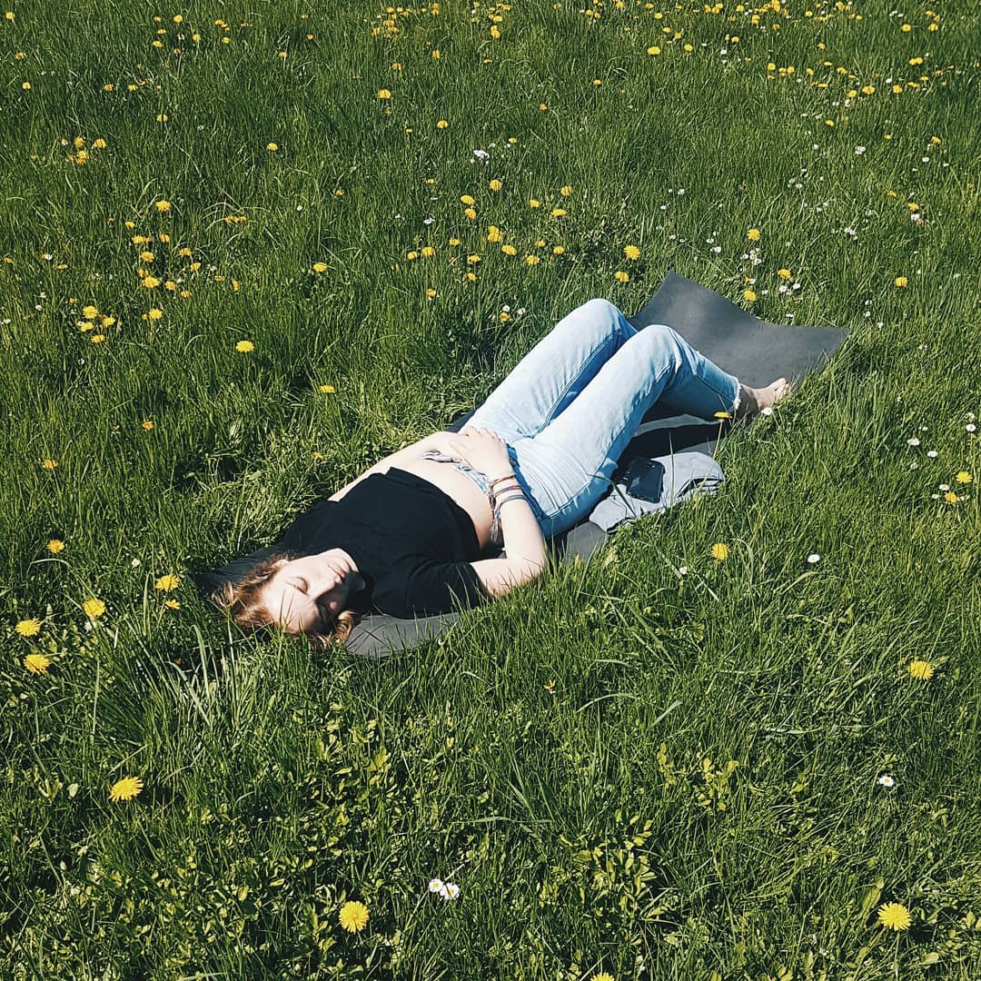 Young woman relaxing in a grassy field with yellow flowers, wearing a black top and jeans, lying on a yoga mat on a sunny day.