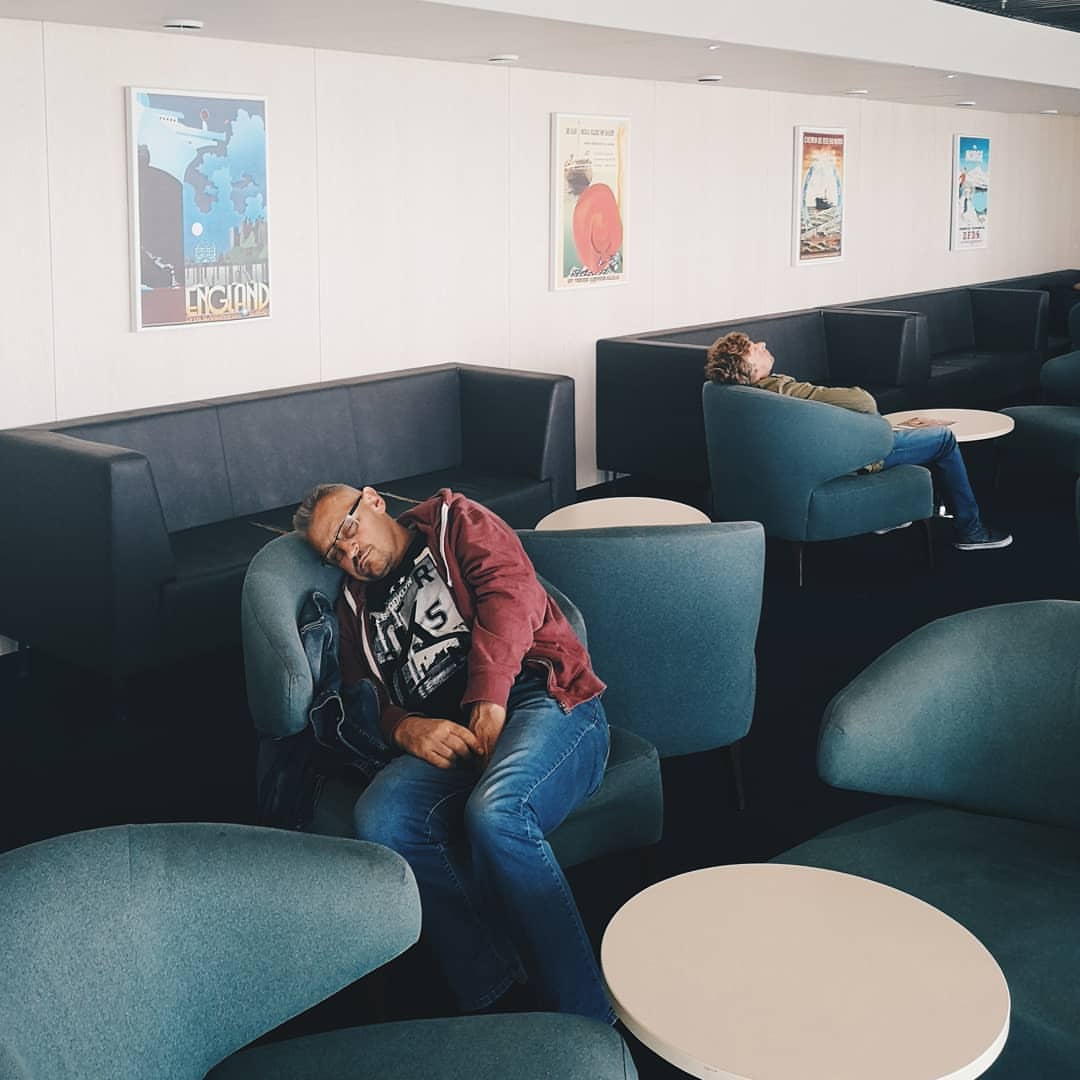 Adult male asleep in a blue armchair in an indoor lounge, with another person reclining in the background and retro posters on the wall.