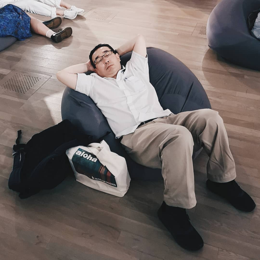 Adult male relaxing on a beanbag chair in an indoor lounge area with a tote bag and backpack nearby, casual wear, eye-level angle.